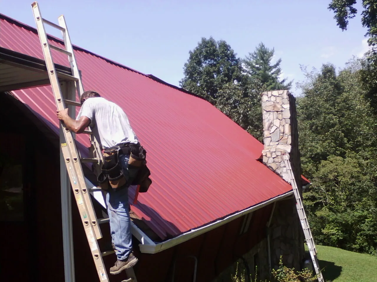 Roofer on a ladder beside a red metal roof and stone chimney — Soffit & Fascia Repair by Roofing Brothers in Warrensburg